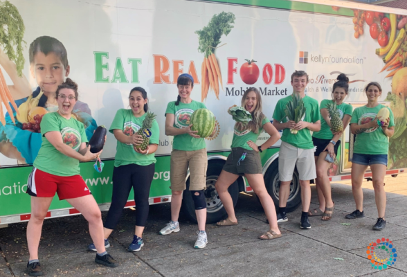 Dr. Meagan Grega with a group of volunteers all standing in front of a sign that says eat real food and each person is holding vegetables and fruit